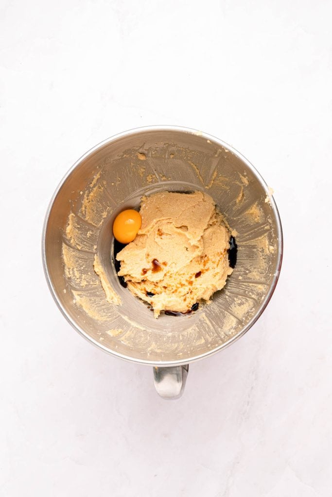 A metal mixing bowl containing cookie dough with one whole egg added on top, viewed from above on a white surface.