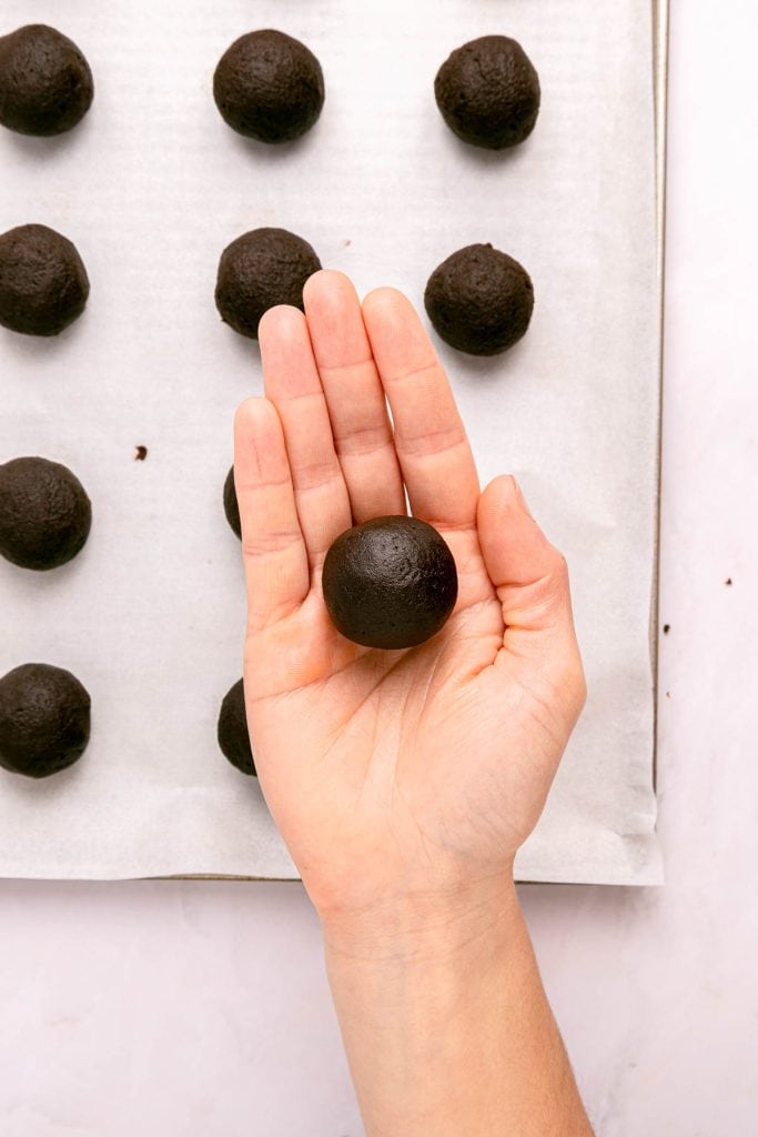 A hand holding a round ball of chocolate dough above a baking sheet lined with evenly spaced dough balls.