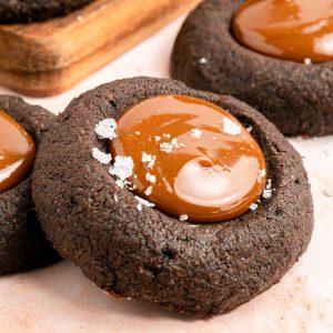 Close-up of a chocolate thumbprint cookie filled with caramel and sprinkled with flaky sea salt, with more cookies in the background.