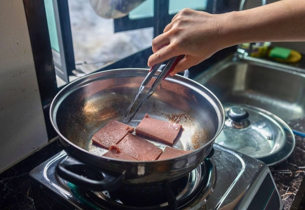 A hand uses tongs to cook four slices of luncheon meat in a stainless steel frying pan on a stovetop near a sink.
