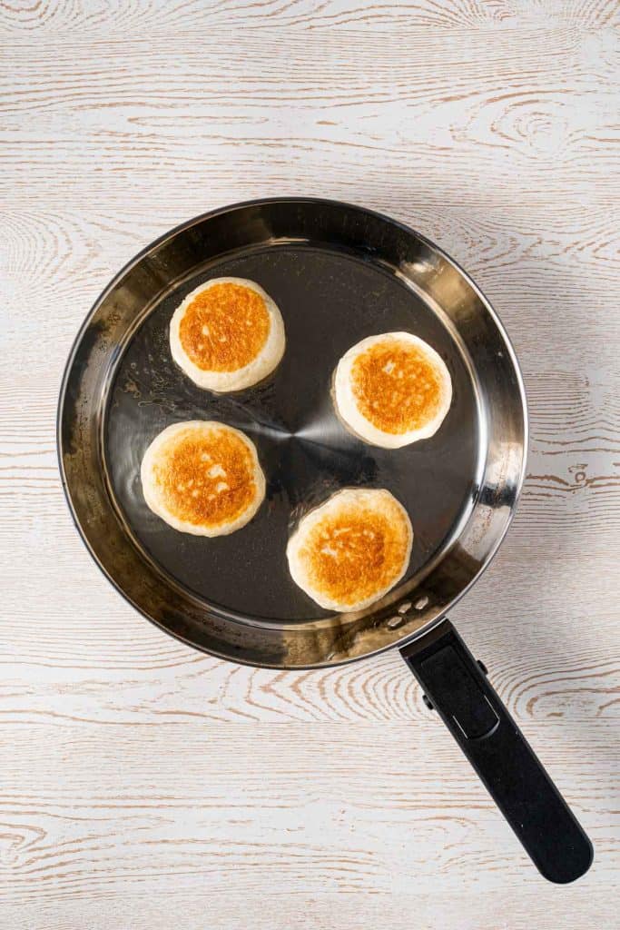 Four round biscuits are being cooked and browned in a stainless steel frying pan on a light wooden surface.