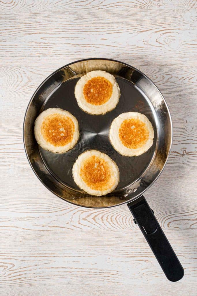 Four round, golden-brown English muffins are cooking in a stainless steel frying pan on a light wooden surface.