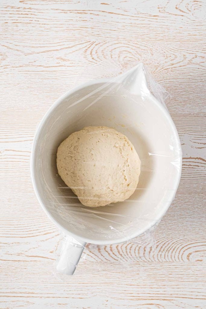 A bowl of dough covered with plastic wrap sits on a light wooden surface.