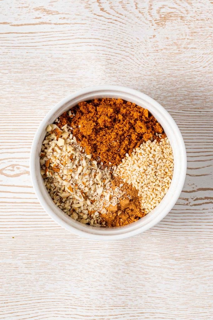 A white bowl containing chopped nuts, brown sugar, sesame seeds, and ground cinnamon placed on a light wooden surface.