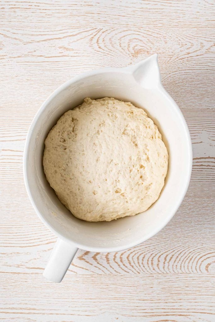 A white mixing bowl containing risen bread dough on a light wood surface.
