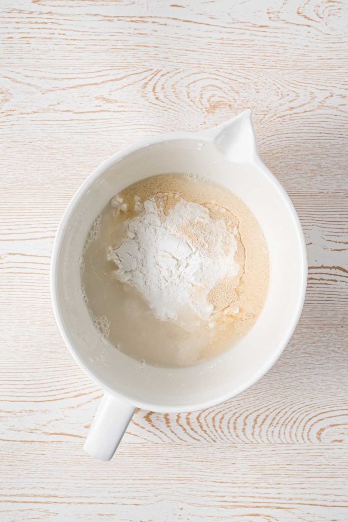 A white mixing bowl containing flour, dry yeast, and water on a light wood surface, viewed from above.