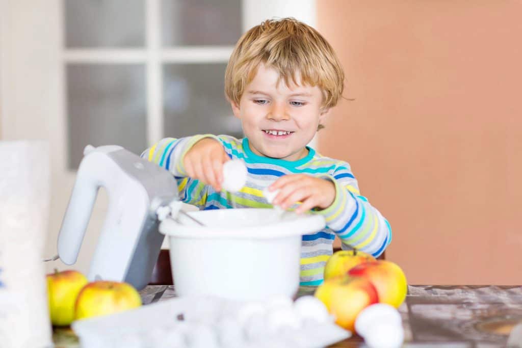 Young child cracking eggs into a mixing bowl on a kitchen table, surrounded by apples, a mixer, and egg carton.