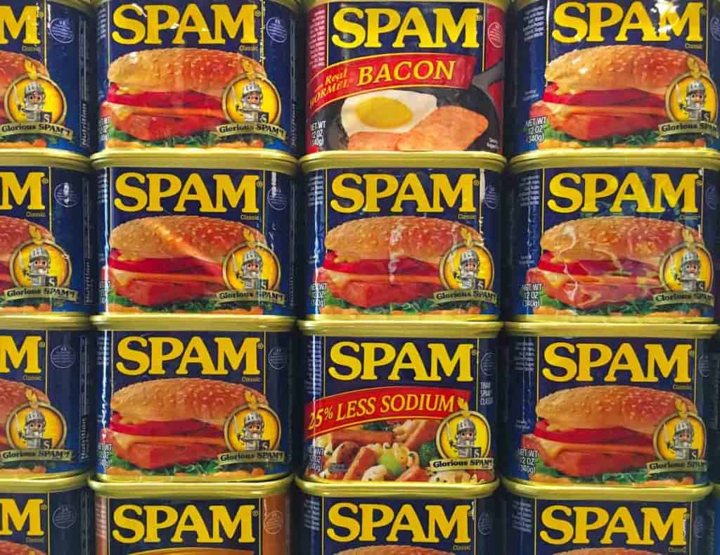 Stacked cans of Spam, including original, bacon, and 25% less sodium varieties, displayed in a grid pattern.