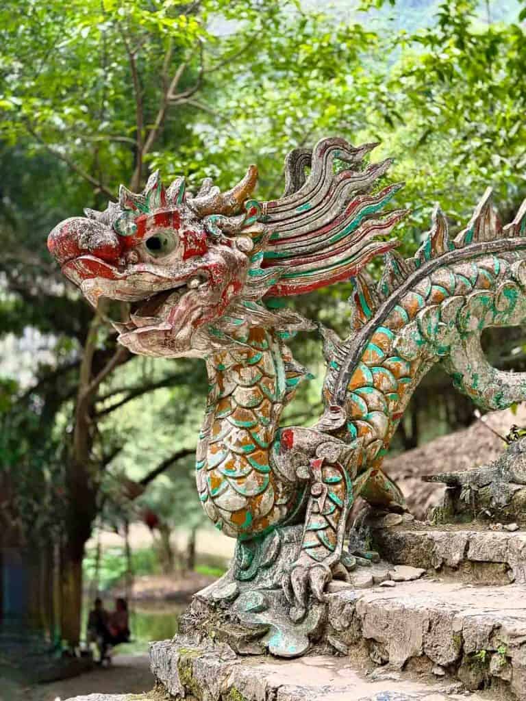 A weathered, colorful dragon sculpture made of stone is positioned outdoors on stone steps, surrounded by trees and greenery.