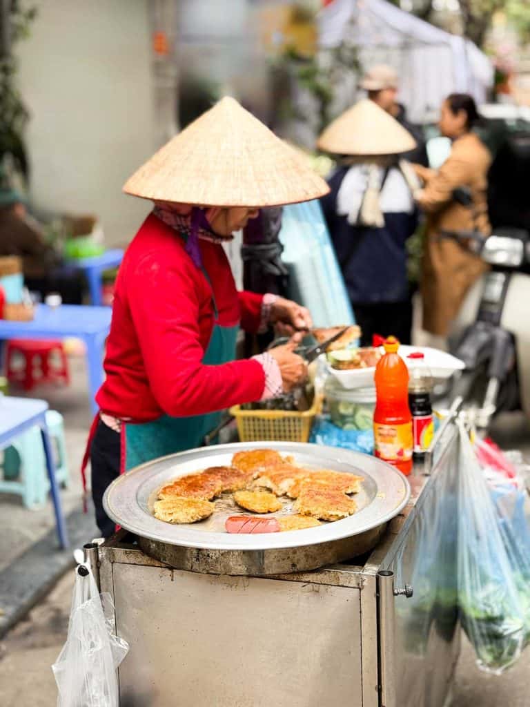 A street vendor wearing a conical hat prepares and sells fried food at an outdoor stall, with cooking supplies and plastic stools visible nearby.