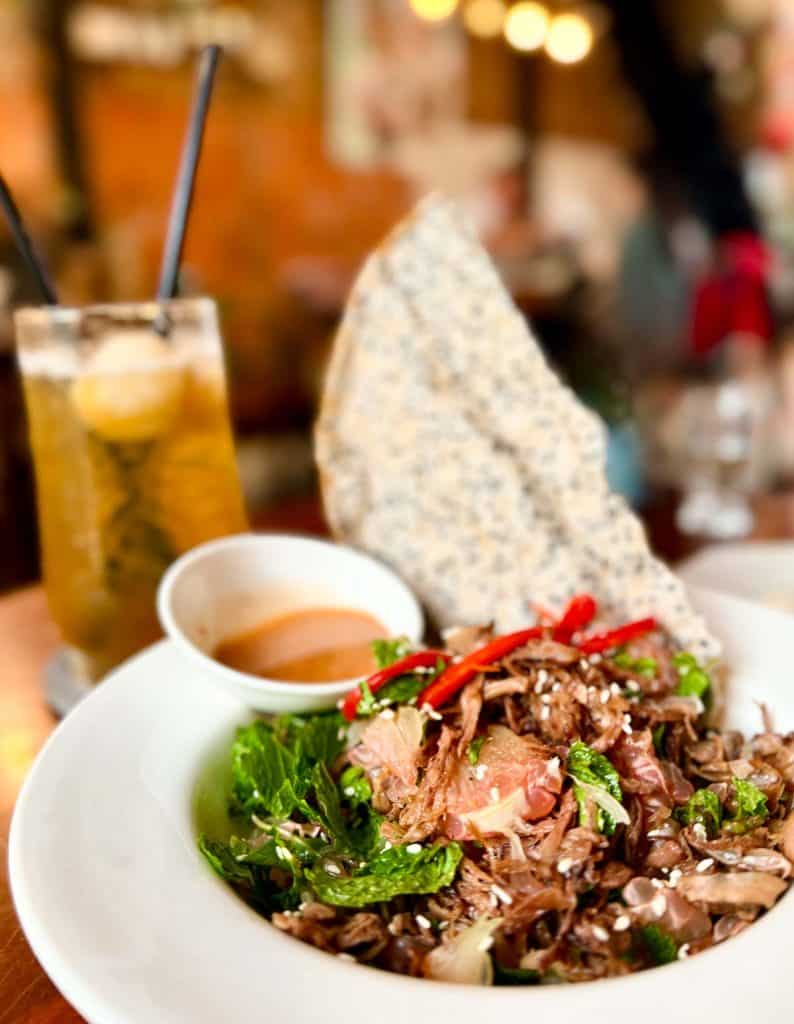 A white bowl with a meat and vegetable salad, garnished with sesame seeds and red chili, served with a dipping sauce and a sesame crisp; iced drink with lemon slices in the background.