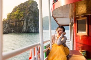 A woman with sunglasses sits on a boat wrapped in a yellow blanket, with water and rocky cliffs visible in the background.