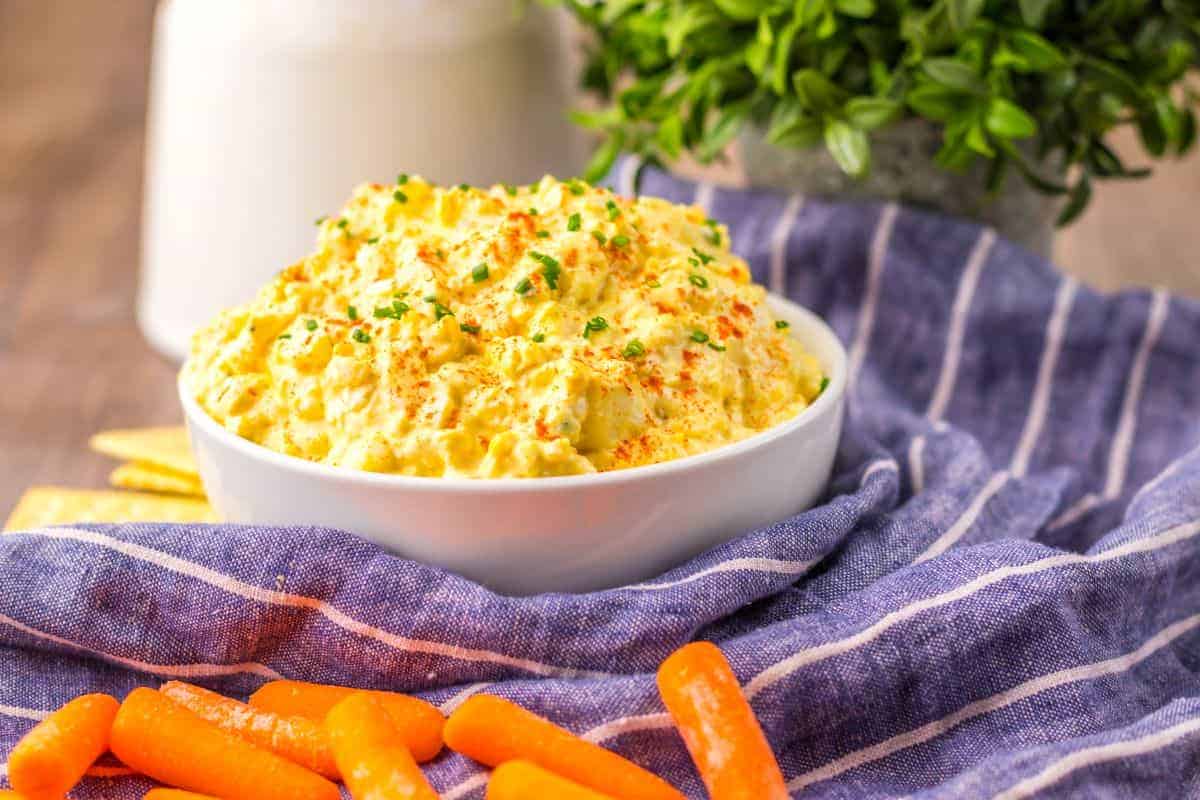 A bowl of deviled egg dip garnished with chives and paprika, surrounded by baby carrots on a striped cloth.