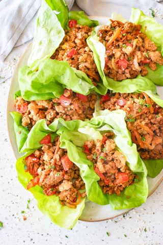 A plate with six lettuce wraps filled with seasoned ground meat, diced tomatoes, and vegetables, arranged on a light-colored surface.