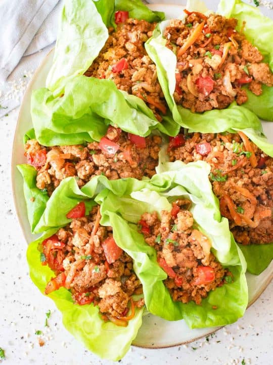 A plate with six lettuce wraps filled with seasoned ground meat, diced tomatoes, and vegetables, arranged on a light-colored surface.