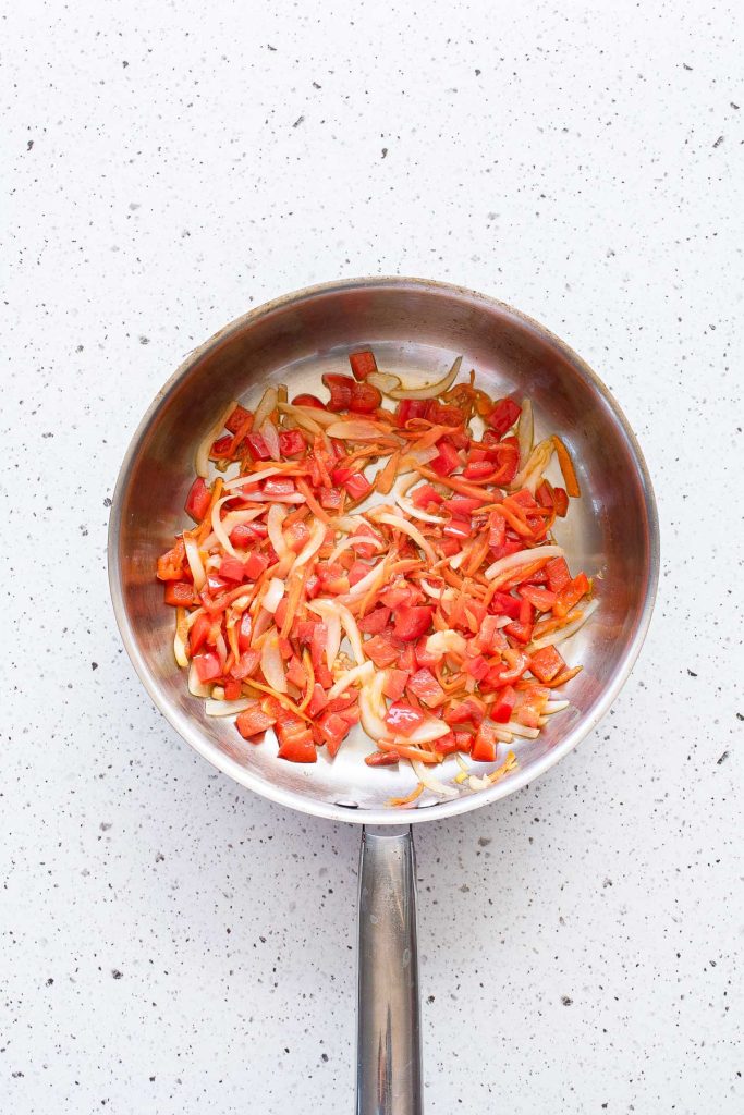 A stainless steel pan with sautéed onions and red bell peppers on a white speckled countertop.