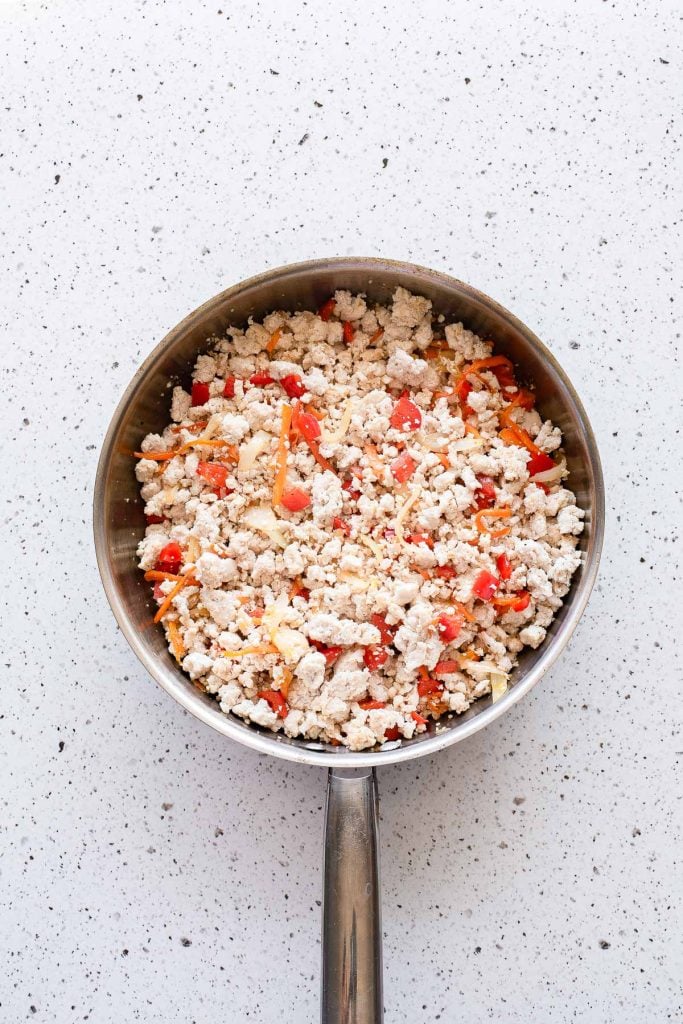 A stainless steel pan on a speckled countertop filled with scrambled tofu, red bell peppers, and grated carrots.