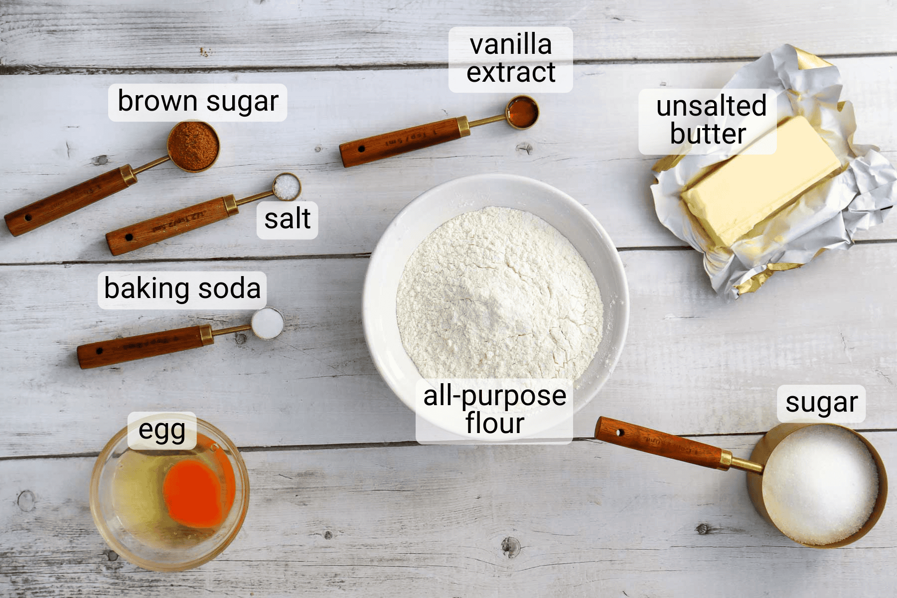 Baking ingredients on a white wooden surface: brown sugar, salt, baking soda, egg, vanilla extract, all-purpose flour, unsalted butter, and sugar, each labeled.