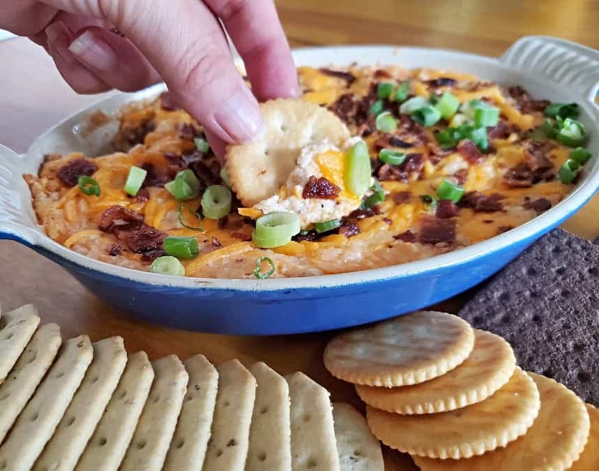 Image shows a hand dipping a cracker into Hot Bacon Cheddar Dip in a ceramic dish.