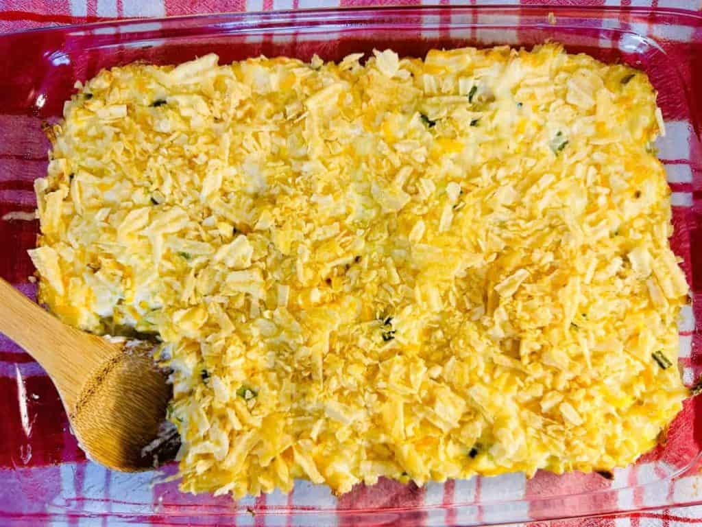 A glass baking dish with a rectangular casserole topped with crushed crackers or chips; a wooden spoon rests in a missing corner serving.