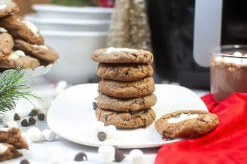 A stack of five chocolate chip cookies with marshmallows sits on a white plate, surrounded by chocolate chips, marshmallows, and a red cloth.