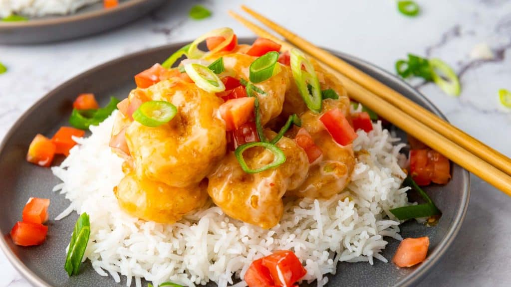 A plate of white rice topped with battered shrimp in a creamy orange sauce, garnished with chopped tomatoes and green onions, with chopsticks on the side.
