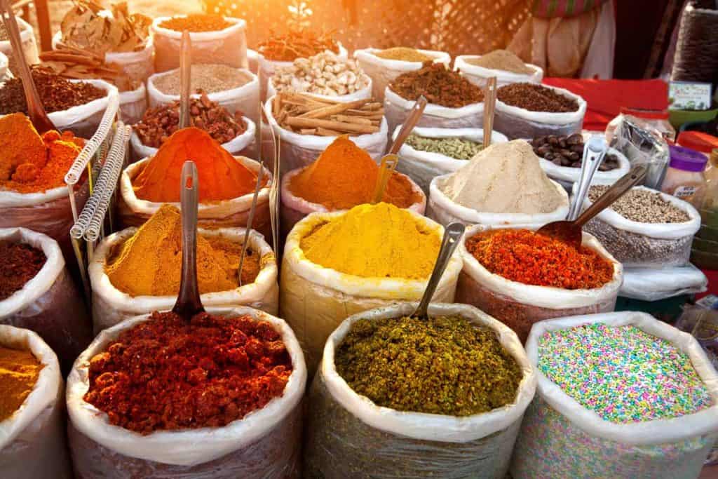Colorful piles of various spices and herbs displayed in large sacks at an outdoor market, each with a metal scoop.
