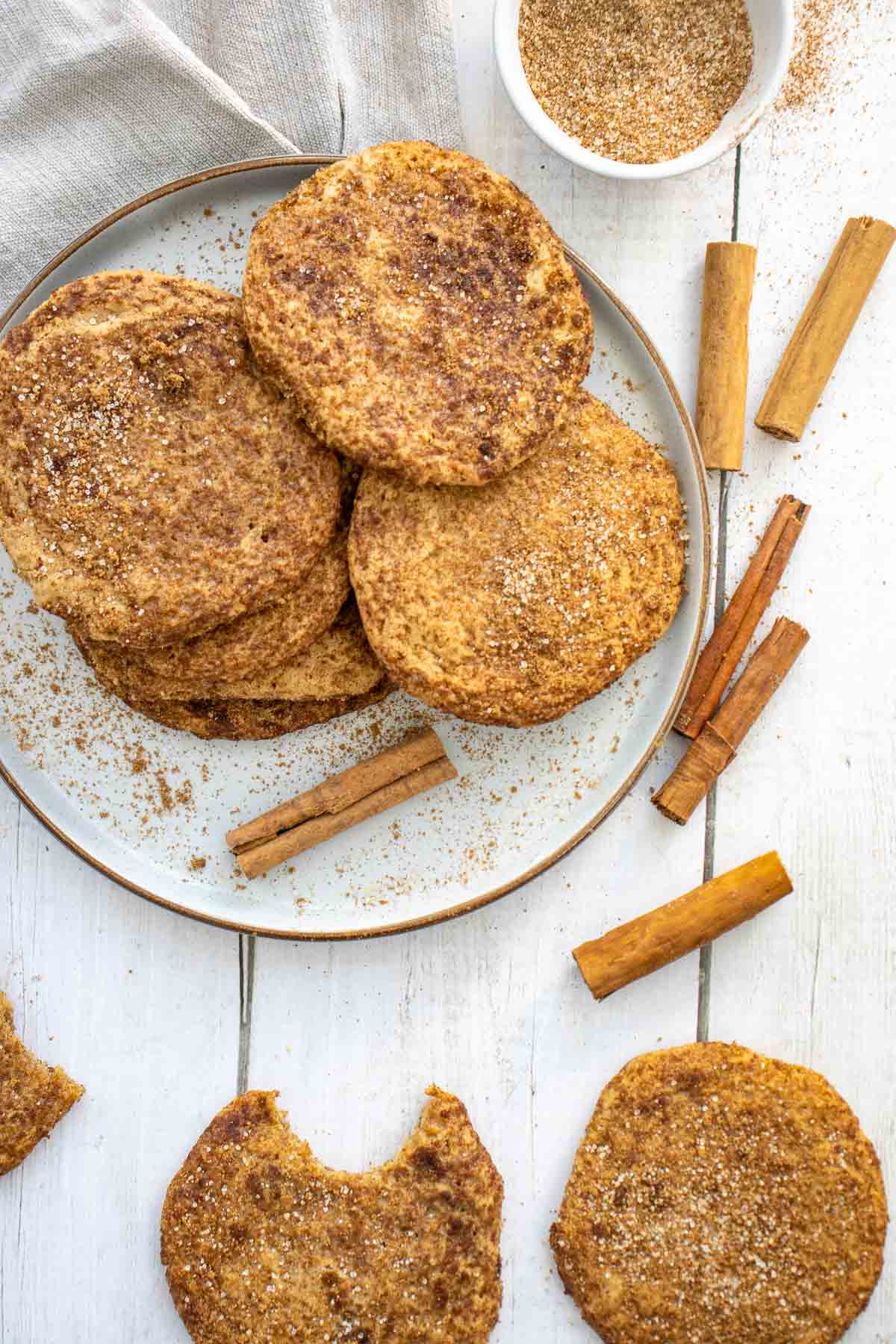 A plate of large snickerdoodle cookies dusted with cinnamon sugar, surrounded by cinnamon sticks on a white wooden surface; one cookie has a bite taken out.