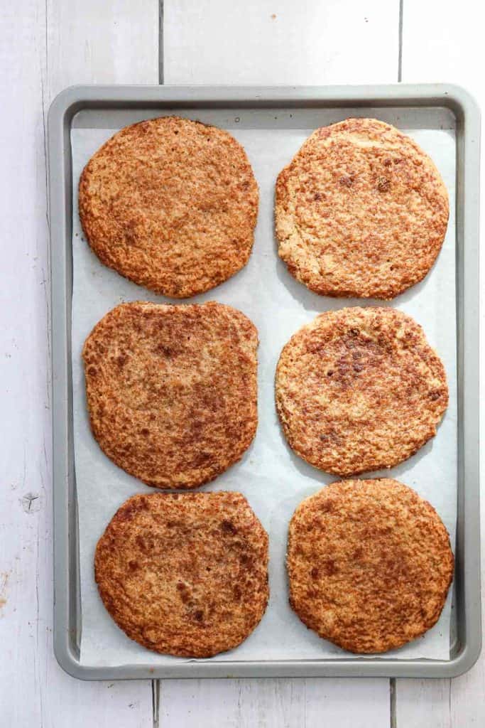 Six round, brown cauliflower sandwich thins are arranged on a parchment-lined baking sheet on a white wooden surface.