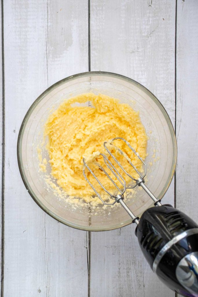 A glass bowl with creamed yellow mixture and an electric hand mixer on a white wooden surface.