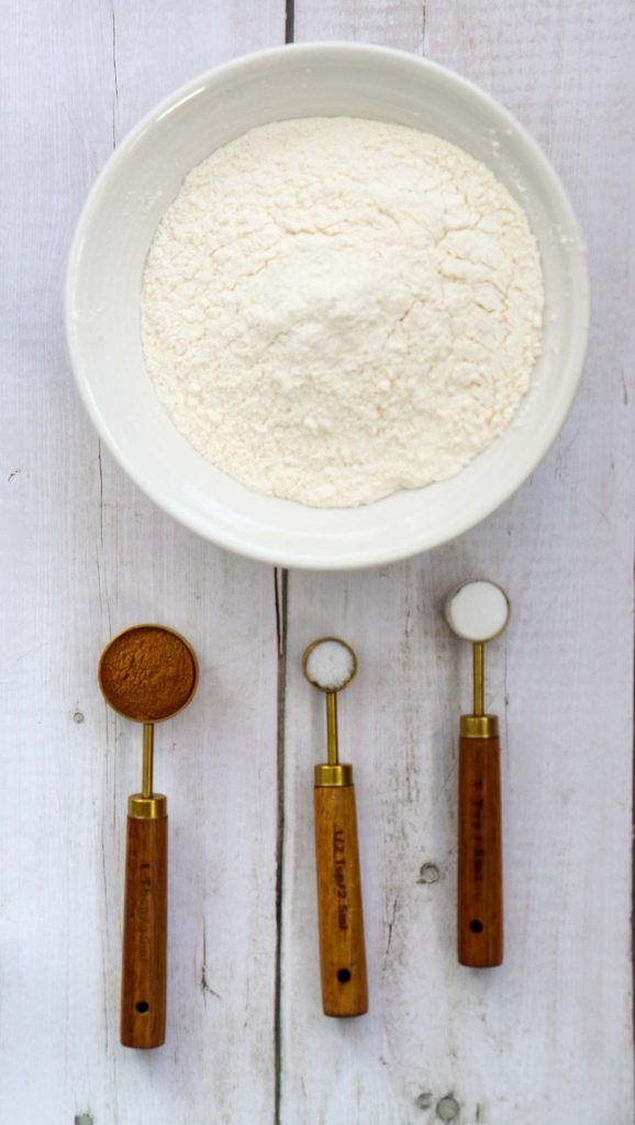 A white bowl filled with flour is placed above three measuring spoons containing cinnamon, baking soda, and salt on a white wooden surface.