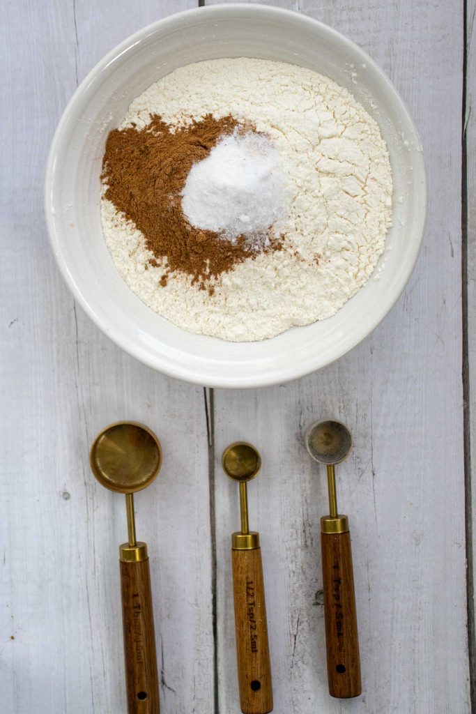 A white bowl with flour, cinnamon, and baking powder on a white wooden surface, with three measuring spoons placed below the bowl.