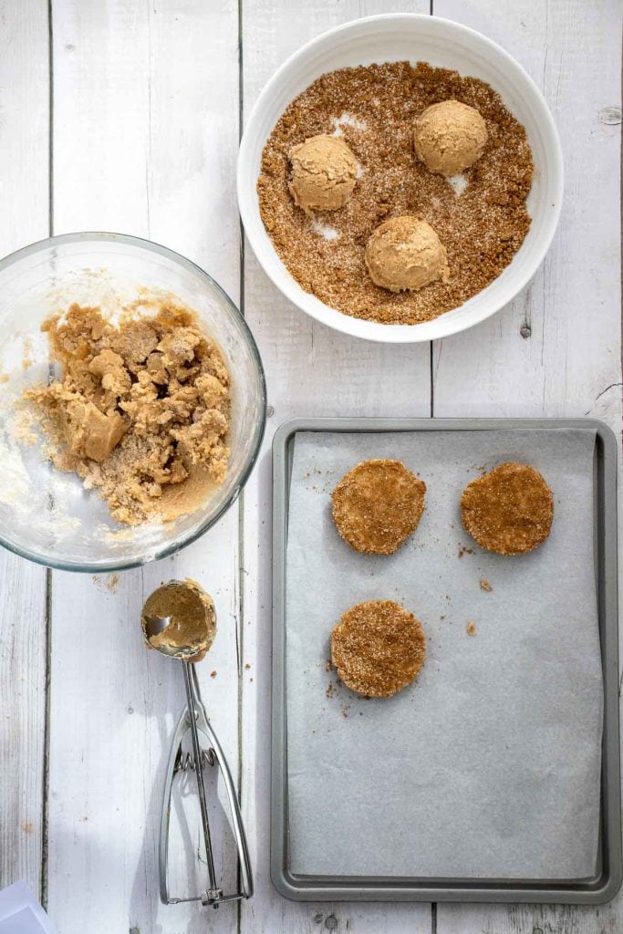 A baking tray with three unbaked cookie dough balls covered in sugar, a bowl of cookie dough, a bowl of sugar, and a cookie scoop on a white wooden surface.