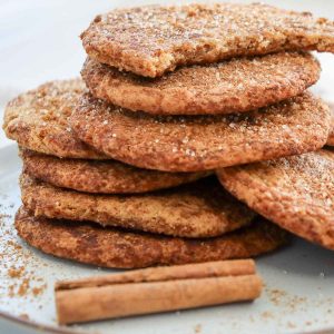 A stack of snickerdoodle cookies on a plate, sprinkled with cinnamon sugar, with two cinnamon sticks in the foreground.