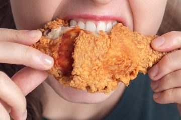 A person bites into a piece of crispy fried chicken, holding it with both hands.