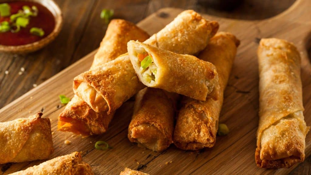 A pile of golden-brown fried egg rolls on a wooden board, with one cut open to show a vegetable filling.