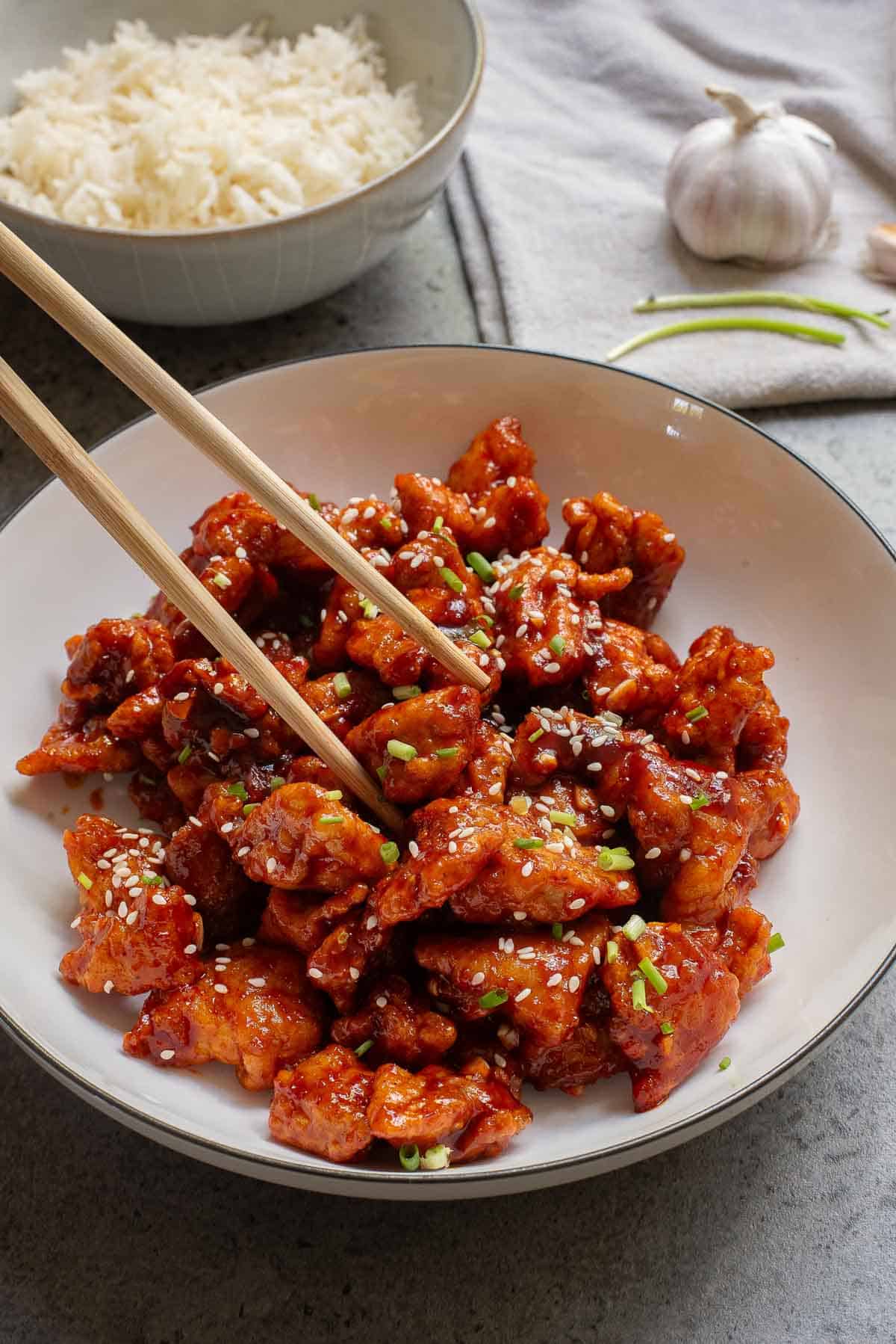 A bowl of saucy, sesame-sprinkled chicken pieces being picked up with chopsticks; a bowl of white rice and garlic are in the background.