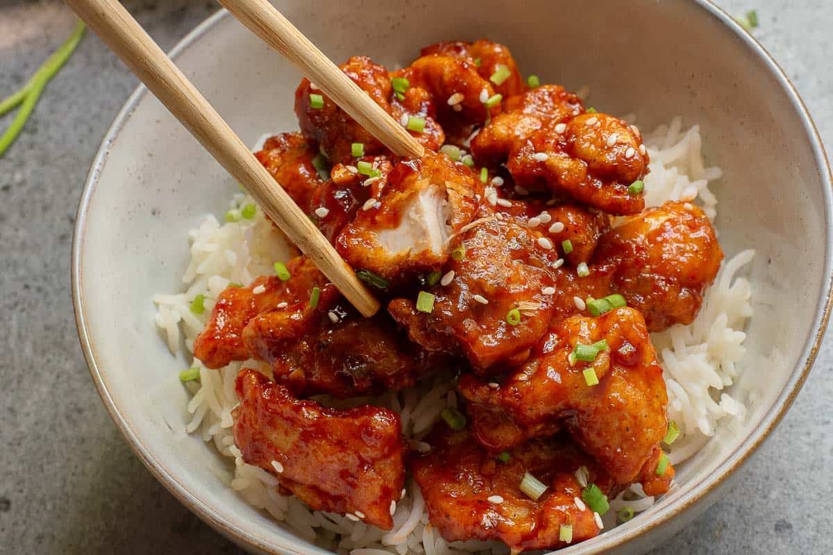 A bowl of white rice topped with crispy, sauced chicken pieces, sprinkled with chopped green onions and sesame seeds, with chopsticks holding one piece.