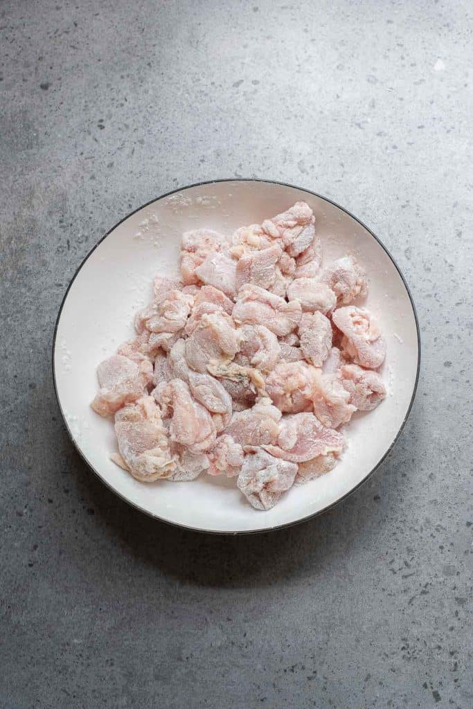 A white plate filled with raw, flour-coated pieces of chicken sits on a gray speckled countertop.