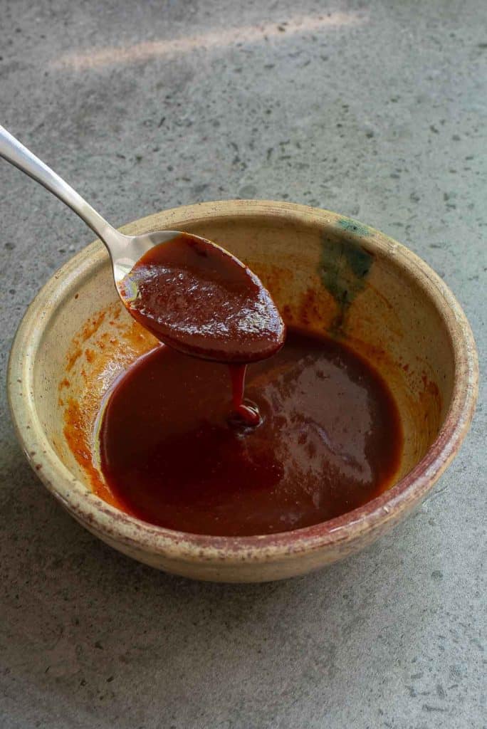 A ceramic bowl filled with reddish-brown sauce, with a spoon holding some of the sauce above the bowl, placed on a gray countertop.