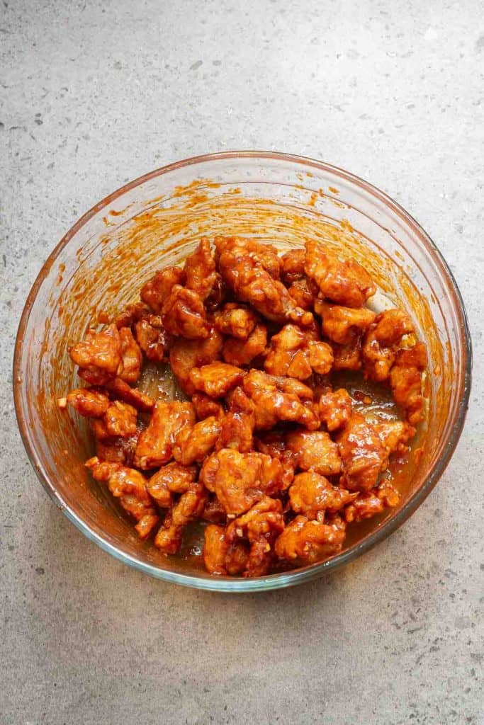 A glass bowl filled with pieces of fried chicken coated in a reddish-orange sauce, placed on a gray countertop.