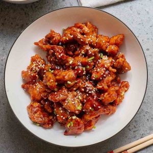 A plate of crispy fried chicken pieces coated in a glossy, reddish-brown sauce, garnished with sesame seeds and chopped green onions.