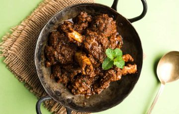 A black metal bowl containing spicy lamb curry garnished with mint leaves, placed on a woven mat beside a gold spoon on a green surface.