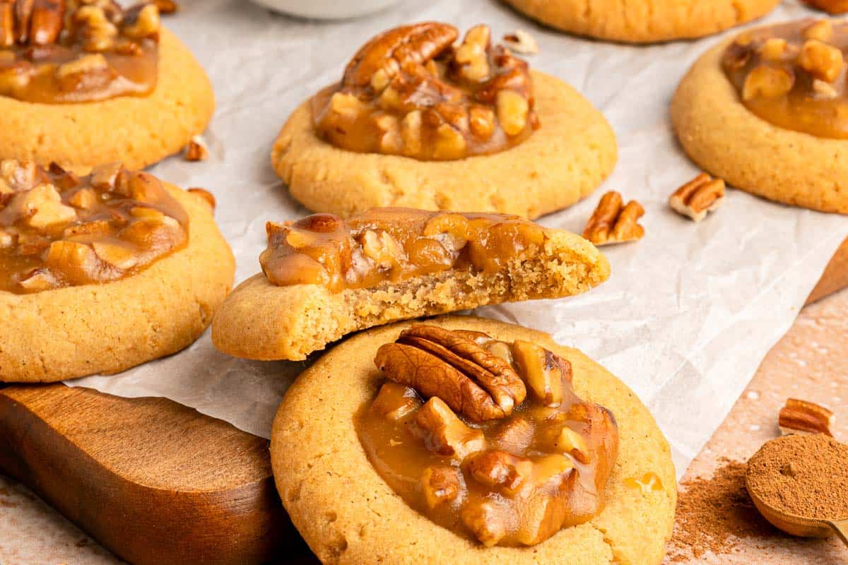 Pecan caramel cookies are arranged on parchment paper, with one cookie broken in half to show the interior. Whole and chopped pecans are visible as toppings.