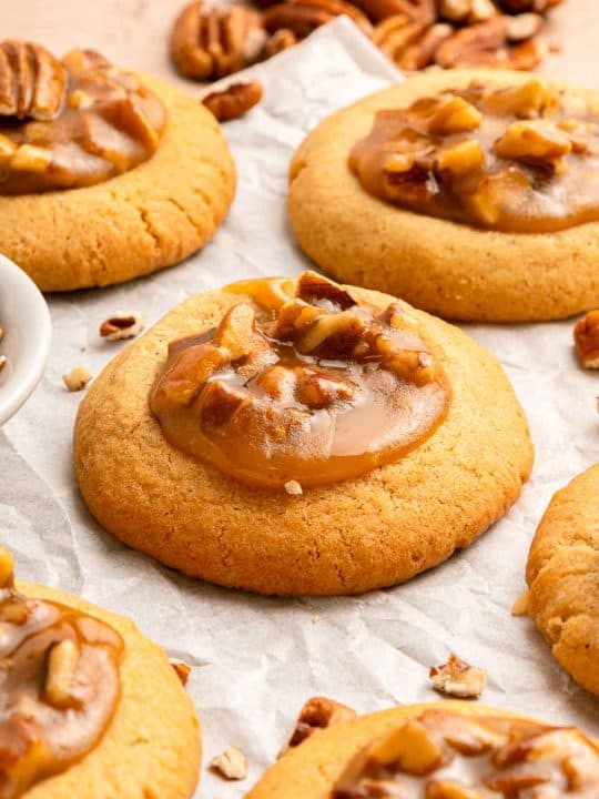 Close-up of several pecan caramel cookies on parchment paper, with chopped pecans scattered around.