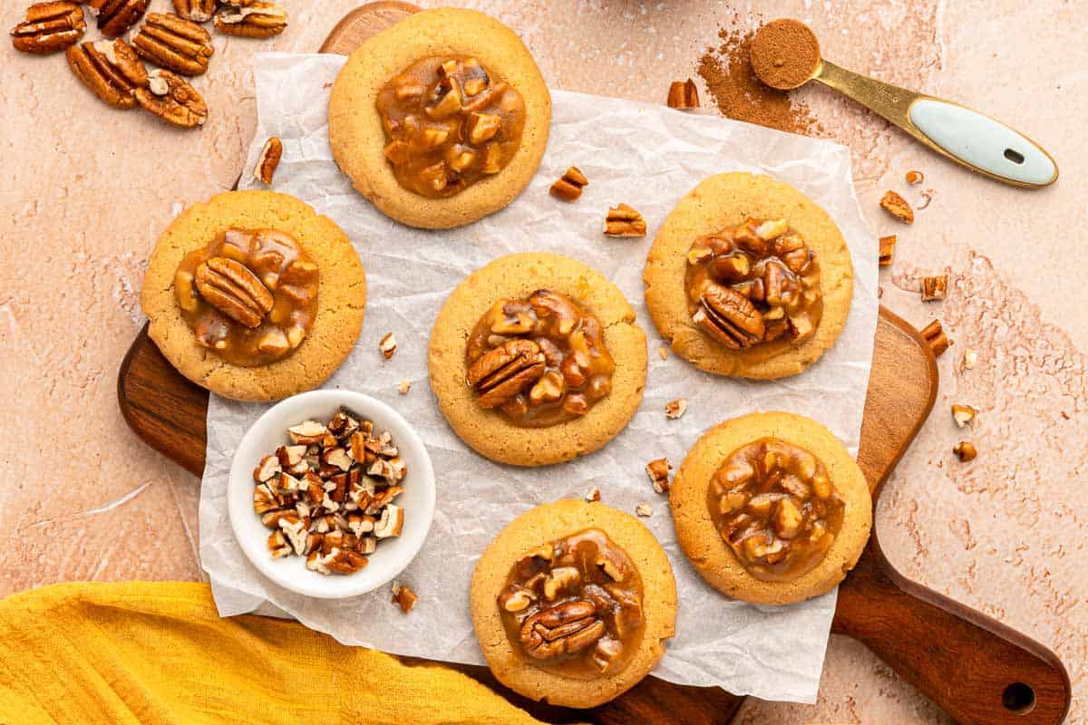 Six pecan cookies with caramel topping are arranged on parchment paper on a wooden board, with a small bowl of chopped pecans nearby and a spoonful of ground cinnamon at the top right.