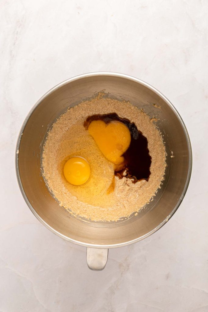 A metal mixing bowl containing creamed butter and sugar, two raw eggs, and vanilla extract on a light countertop.