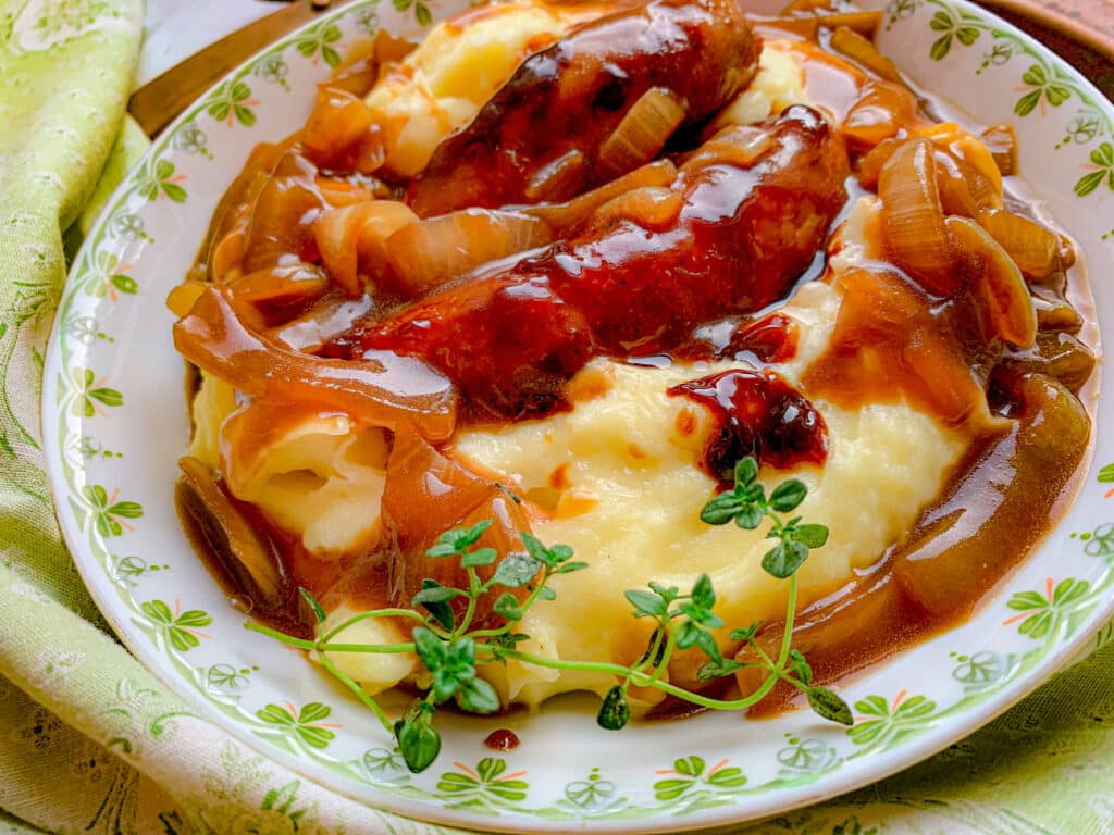 A plate of mashed potatoes topped with sausages and caramelized onions in a rich brown gravy. A sprig of fresh thyme garnishes the dish. The plate sits on a green patterned cloth.