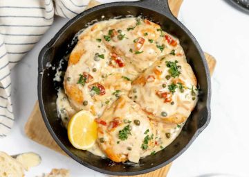 Skillet with four pieces of chicken in creamy sauce with capers, chopped herbs, and sundried tomatoes, garnished with a lemon wedge.