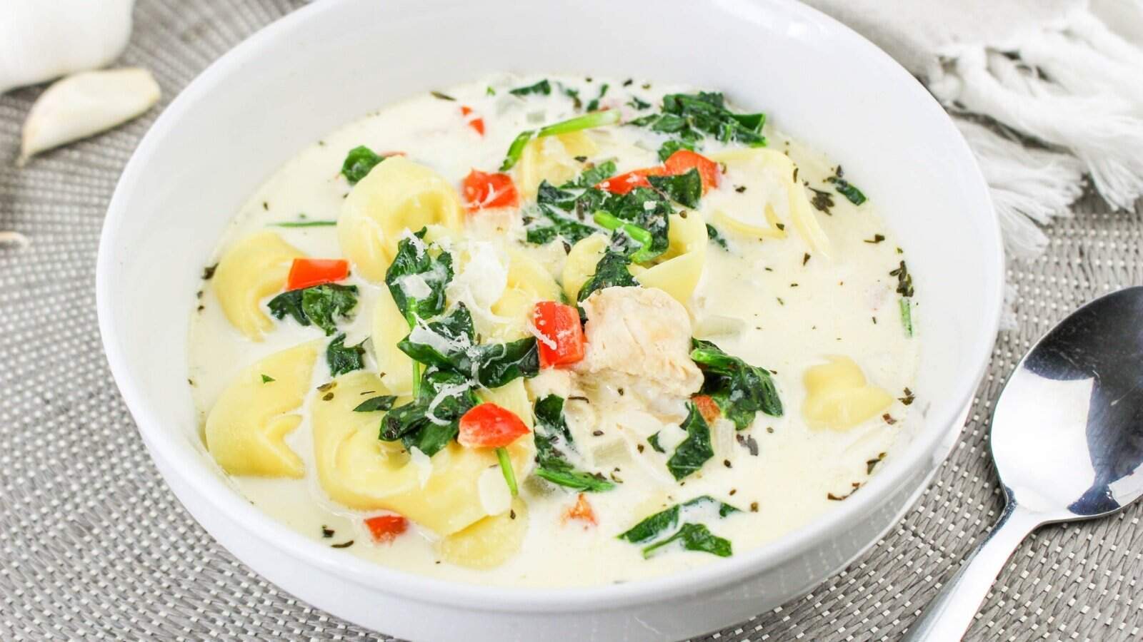 A white bowl filled with creamy tortellini soup, containing spinach, red peppers, and chicken, on a gray textured placemat with a spoon nearby.